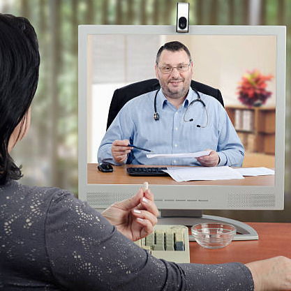 Female patient sits with her back to the camera and looks at virtual doctor in the monitor. Mature woman holds a pill by hand and ready to take it. Middle-aged bearded male doctor with glasses and a stethoscope reviews medical laboratory results with her. There are monitor, keyboard, glass water on wooden desk. Horizontal shot on blurry indoors background.